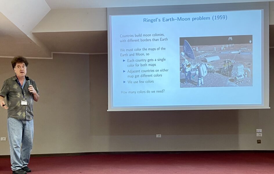 Eppstein stands with a microphone in front of a projector explaining Ringel’s Earth-Moon problem.