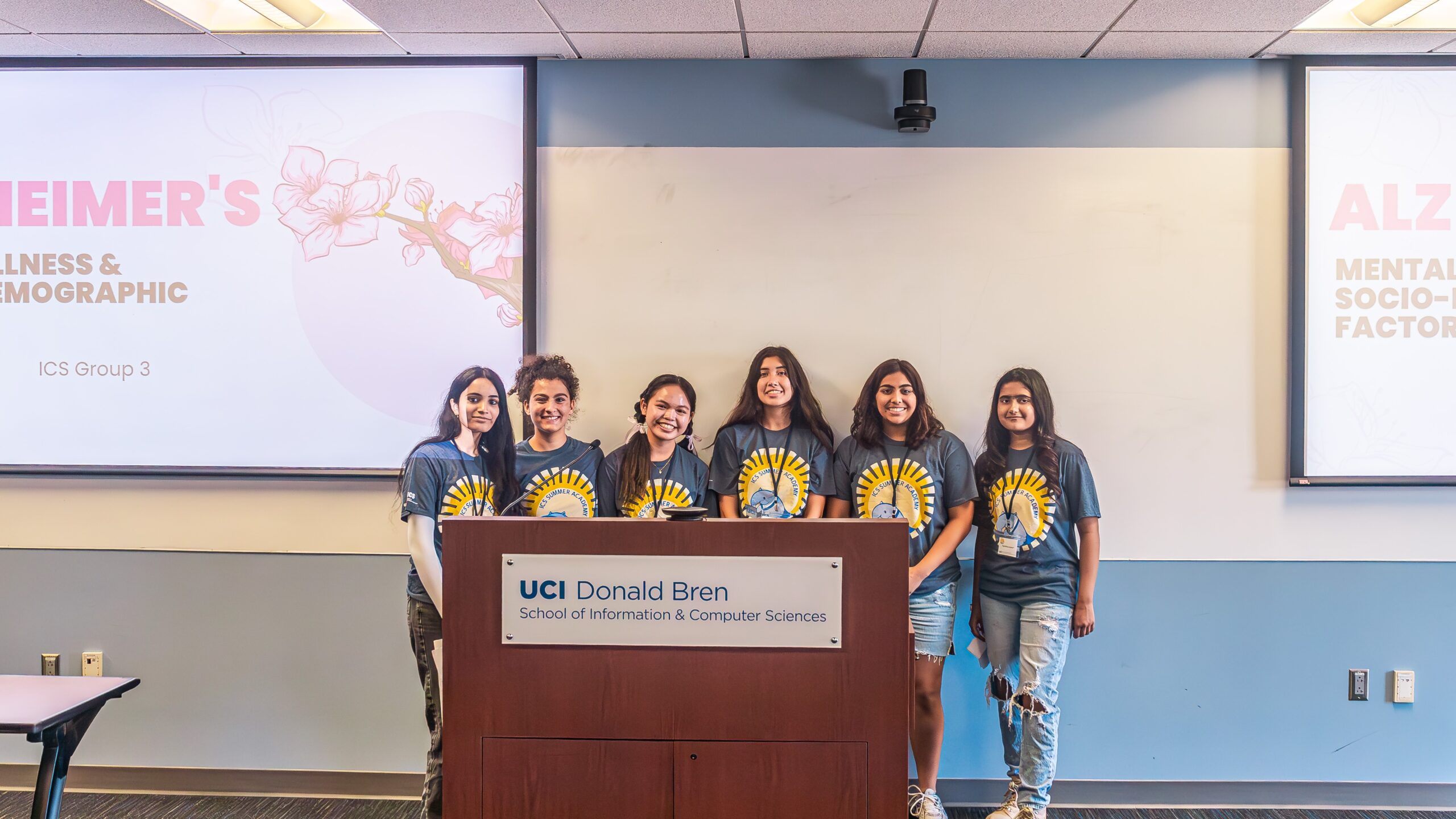 A group of six girls stand behind a podium with their presentation on two screens behind them.
