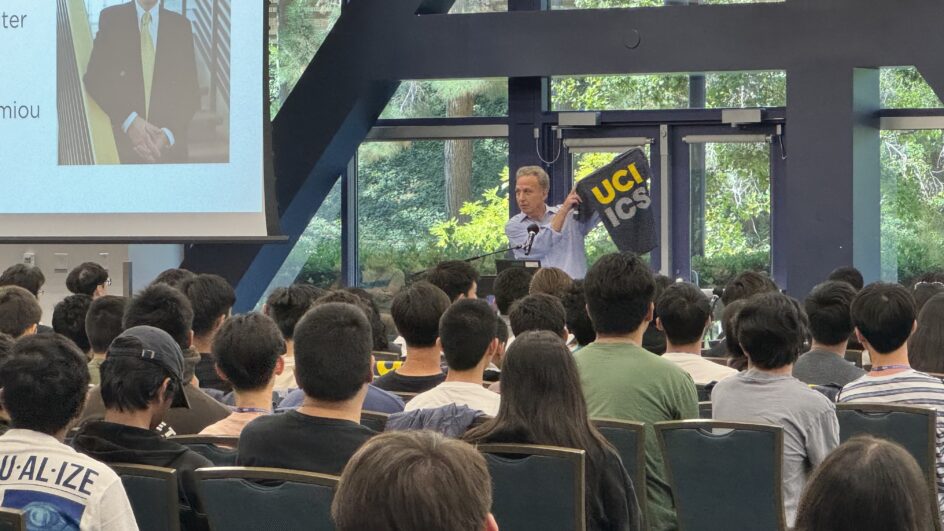 Students sitting in a student center ballroom, with Dean Marios at the front of the room holding up the UCI ICS t-shirt.