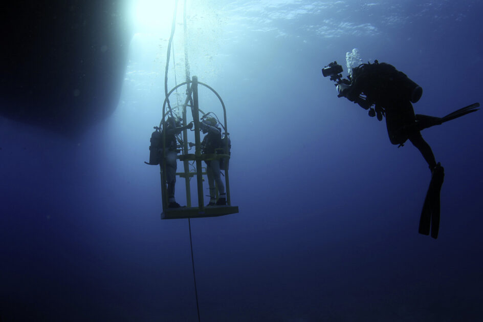 An underwater scene, with Balacy off to the side, taking photos of two other people standing on a small platform.