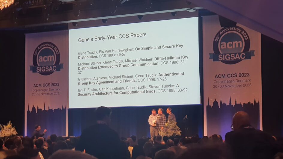 Tsudik appears on stage at the conference, with some of his early year CCS papers appearing on a screen behind him.