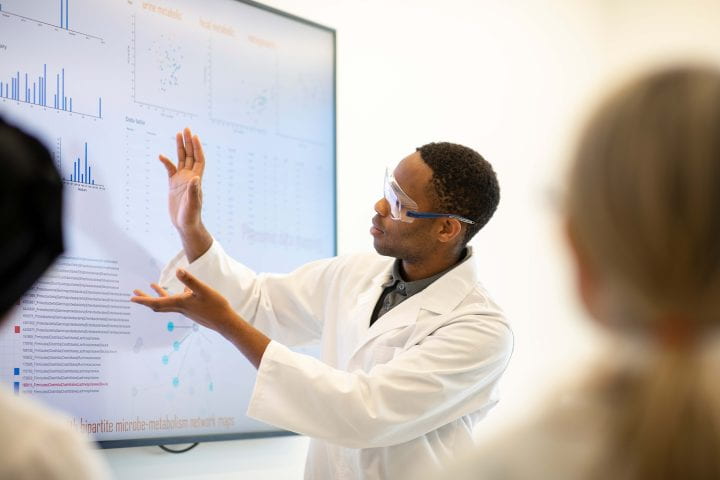 A man with goggles and a white lab coat facing a white board with graphs.