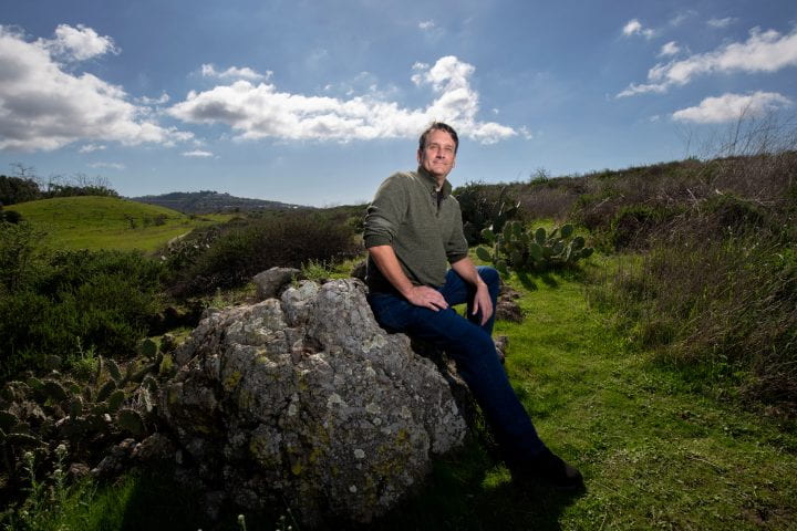 Bill Tomlinson leans against a rock in a field of grass with blue skies in the background