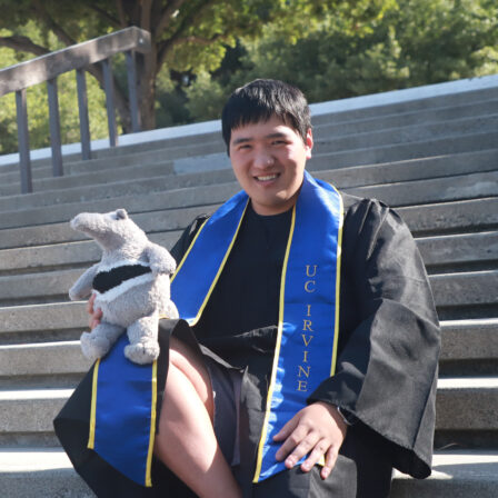 Shengyuan Lu sits on stairs at UCI in graduation gown with an anteater stuffed animal