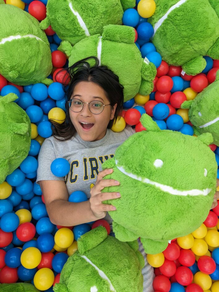 Maya Lee lying on top of colorful balls and plushies of the Android green robot, holding one of the plushies in her hands. 