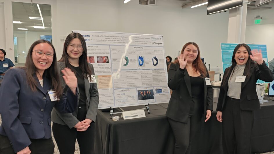 The four students, standing near a poster about their work, smile and wave.