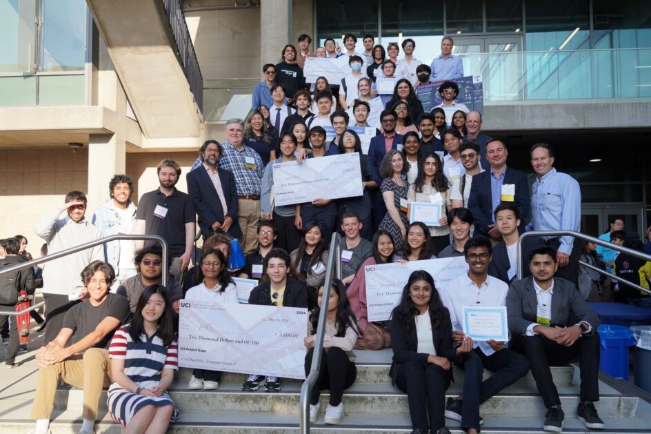 A large group of students, some holding oversized checks, stand together outside a building at UCI