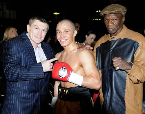 Vieyra stands between Ricky Hatton and Floyd Mayweather Sr., in his boxing shorts, smiling, with his boxing glove in front of his chest.