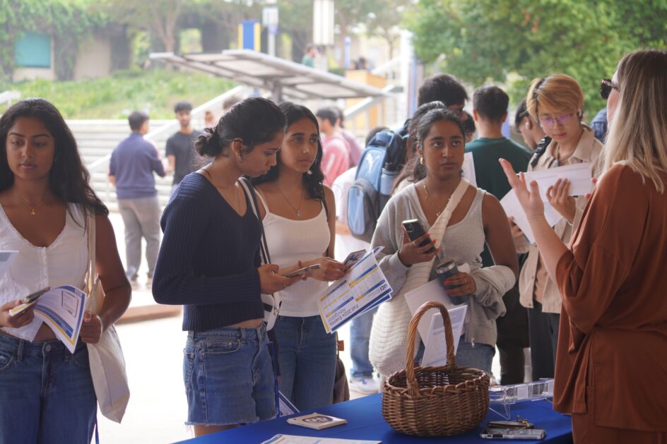 Several students stand at a table, talking to a staff member.