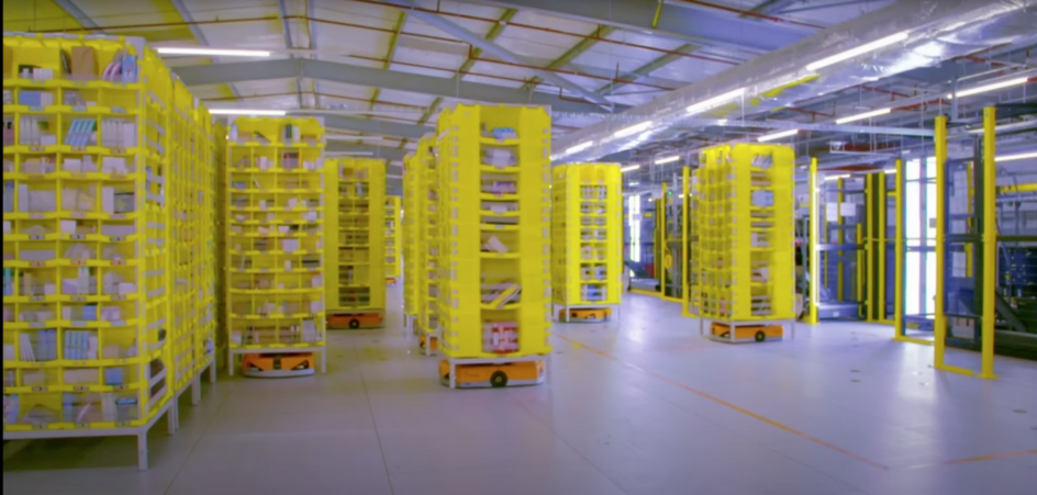 A warehouse with a tall narrow shelves, full of products, with small rectangular robots with wheels sitting underneath some of them.