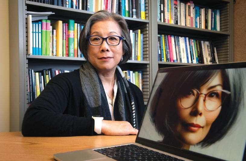 Mimi Ito sits at a desk with a laptop displaying an AI-generated image of her