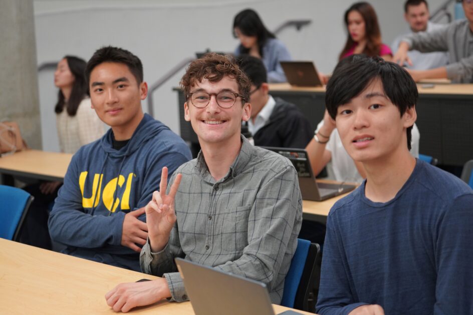 Three male students sit at a table in an auditorium