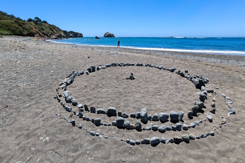 A circle of stones decorates Little River Beach, in Mendocino County, California. Two more partial rings encircle it, with a small cairn of stones at the center. A lone beachwalker in the distance is dwarfed by the perspective; a rocky islet and a line of breaking waves near the horizon meet a blue sky. Farther down the beach, colorful kayaks wait to be taken out under a green bluff.
