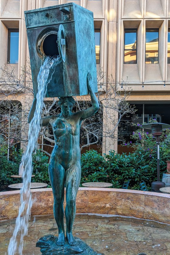 Body of Urban Myth, Brian Goggin, 1997, in the patio of Caffe Riace, Palo Alto. Within a brutalist plaza, a classically-styled bronze statue of a nude woman stands in a pool amid greenery, holding a clothes washing machine over her head. Water pours from its open door.