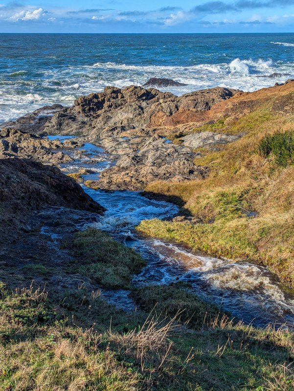 A small stream rushes into the rocky coast of the Pacific Ocean under a cloudy but blue sky at the Point Cabrillo Light Station State Historic Park near Mendocino, California